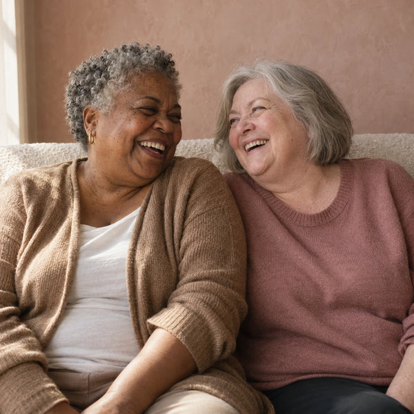 Two women laughing together on a sofa, one in a camel cardigan and one in a dusty rose sweater